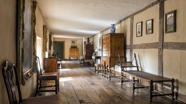 A long, low ceilinged corridor with wide wooden floorboards and half-timbered walls. Antique furniture and paintings line the walls.
